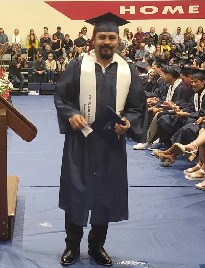 Picture of hispanic man at graduation ceremony, wearing cap and gown and holiding a diploma.