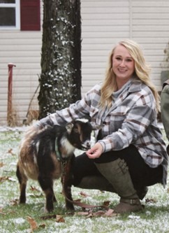 Picture of blond woman with plaid long sleeve shirt bending down and feeding a goat.