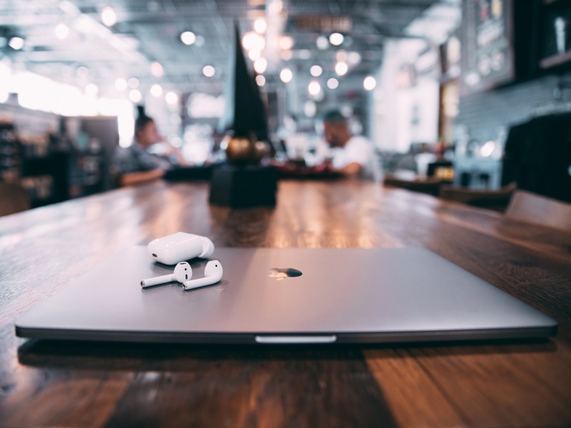 A laptop with the Apple logo and headphones lies on a table with two people in the background