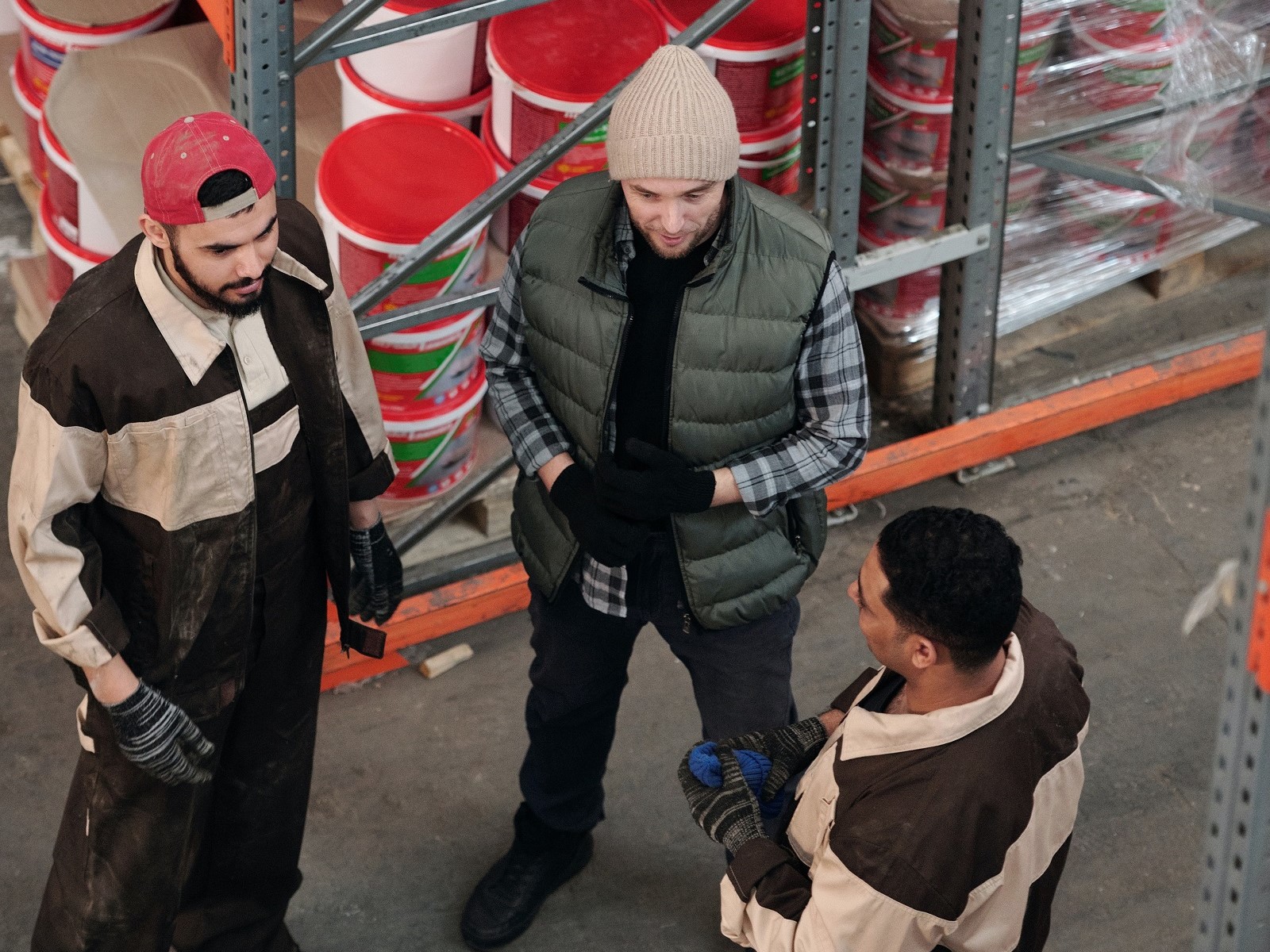 Three men wearing tan and brown jackets talking in warehouse with boxes and orange buckets. 