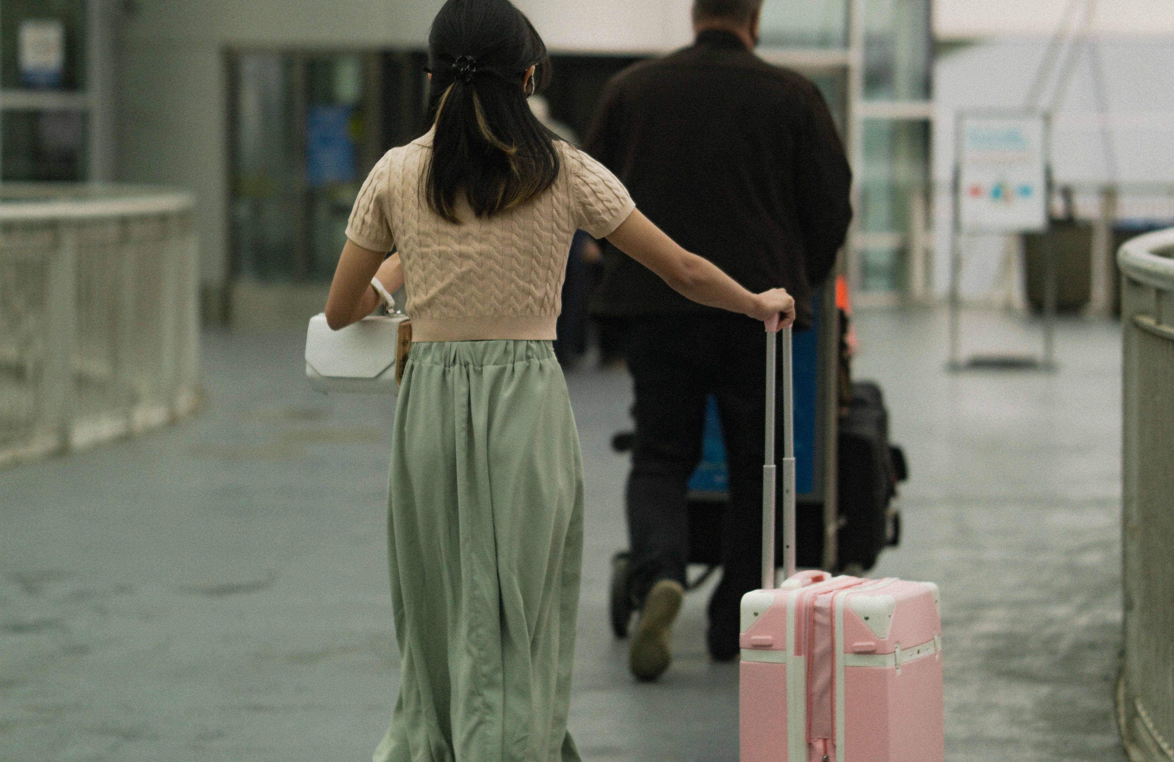 Woman with dark hair in brown T shirt pushing her pink luggage through the airport and man in black long sleeves pushing trolley