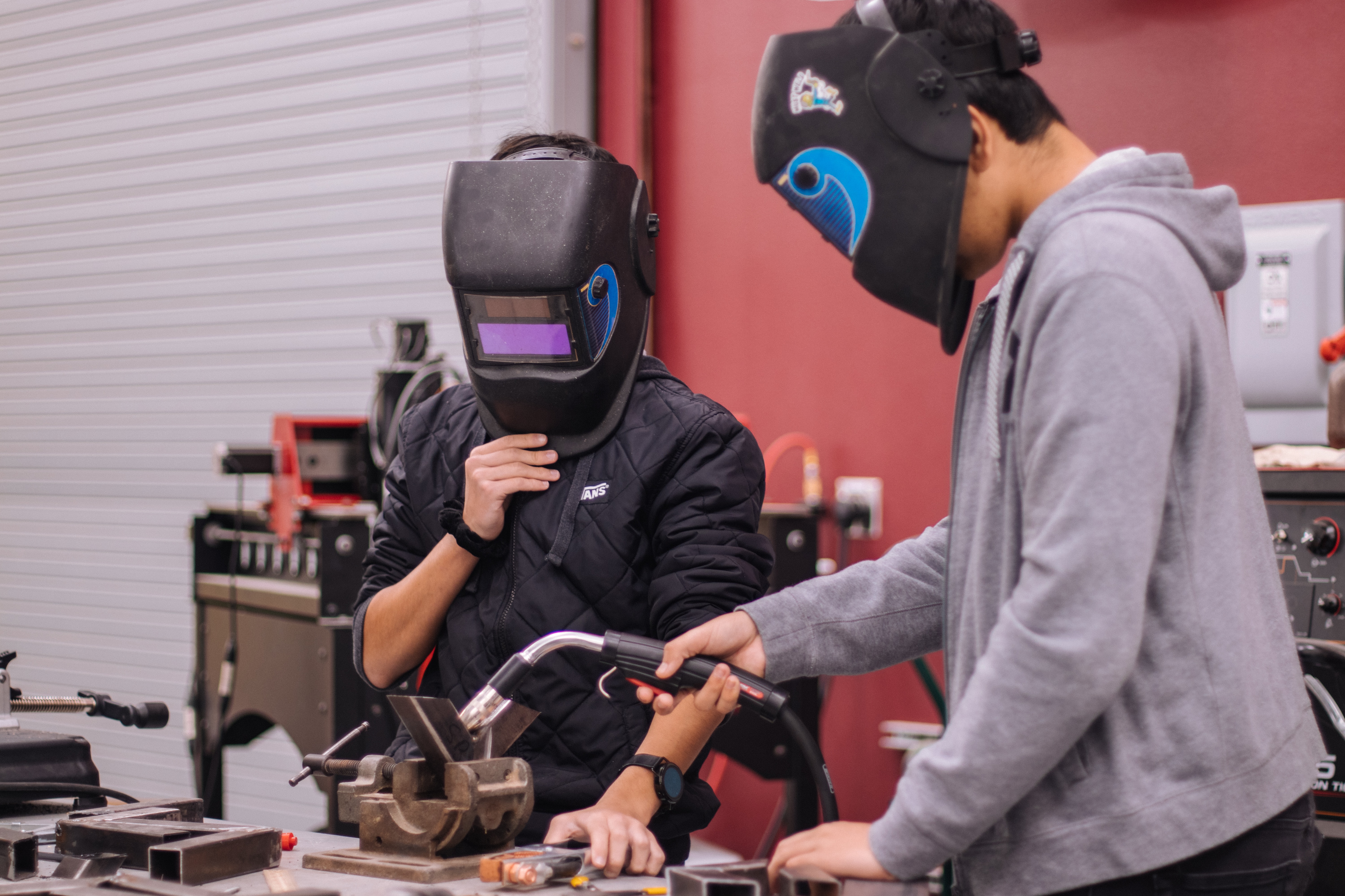 Two kids in welding masks holding a welding gun over a piece of metal