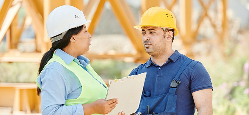 A woman in a white hard hat and green vest holding a clipboard talking to a man in a yellow hard hat and blue shirt