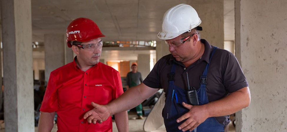 Man in a red shirt and red construction helmet talks to a man with blue overalls and white hat at a construction site