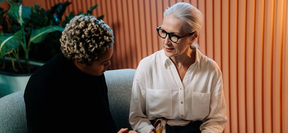 Woman with white hair, black glasses wearing a white shirt talks to a woman with blonde hair in a black shirt sitting on a gray couch