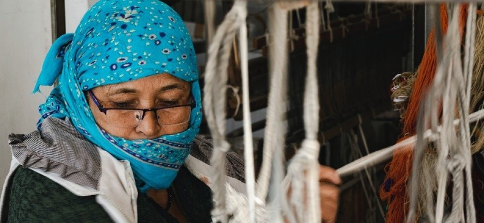 A woman in a blue headscarf wearing a mask and using a hand loom to weave