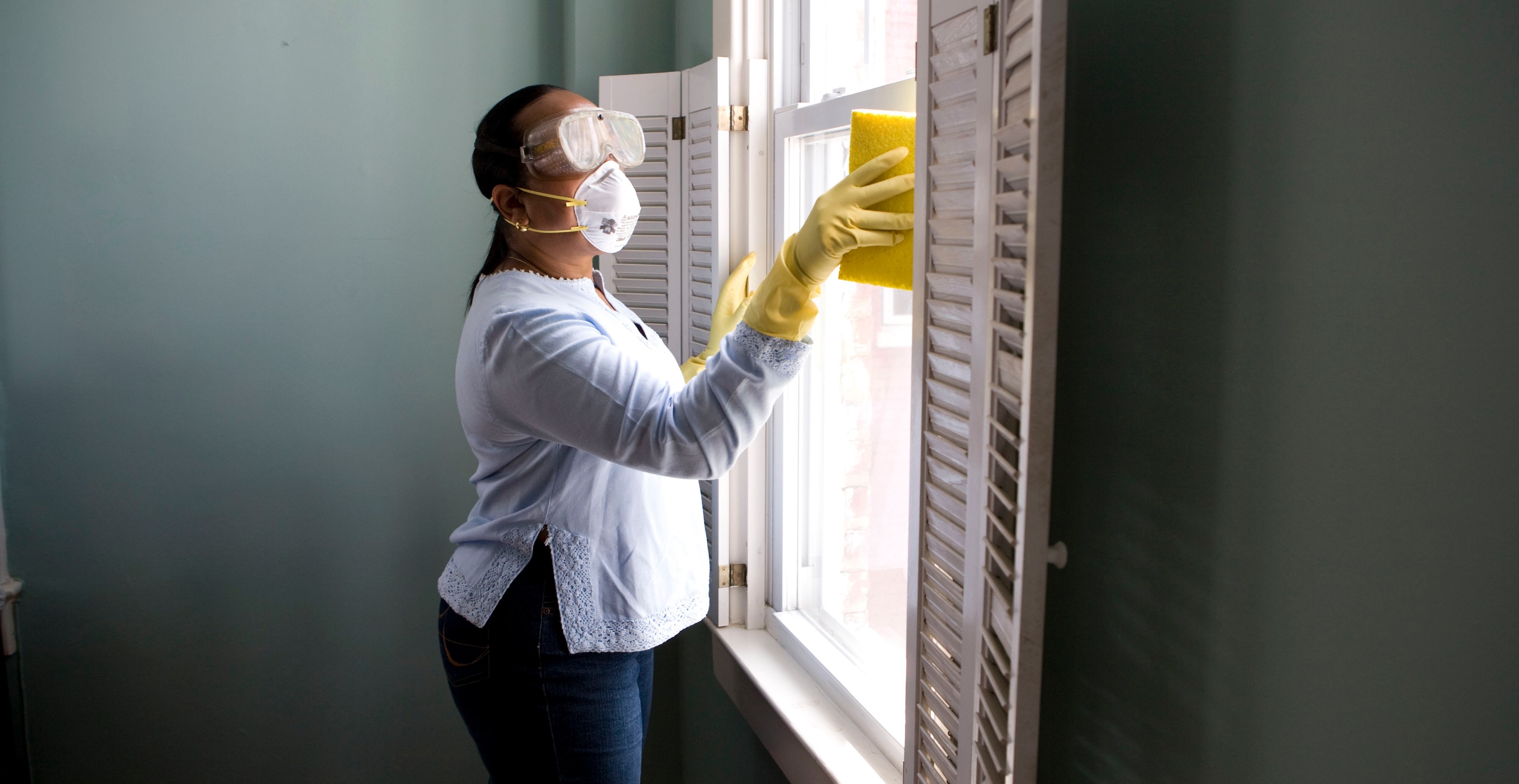Woman in white long sleeve shirt and blue denim jeans cleaning a glass window with a yellow sponge while wearing a mask, goggles and yellow gloves